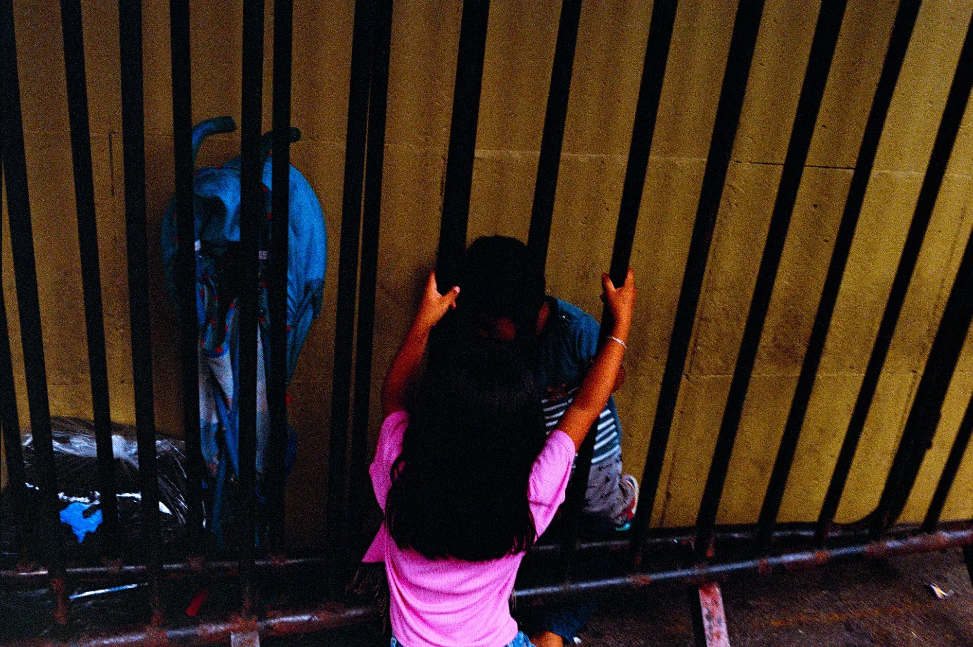 Dos niños se asoman entre los barrotes de una reja frente a un muro amarillo