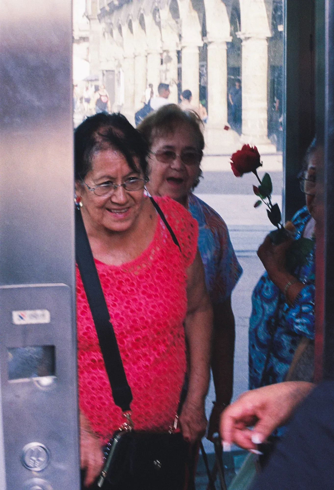 Tres mujeres mayores sonríen al salir por una puerta de cristal; una lleva una rosa roja
