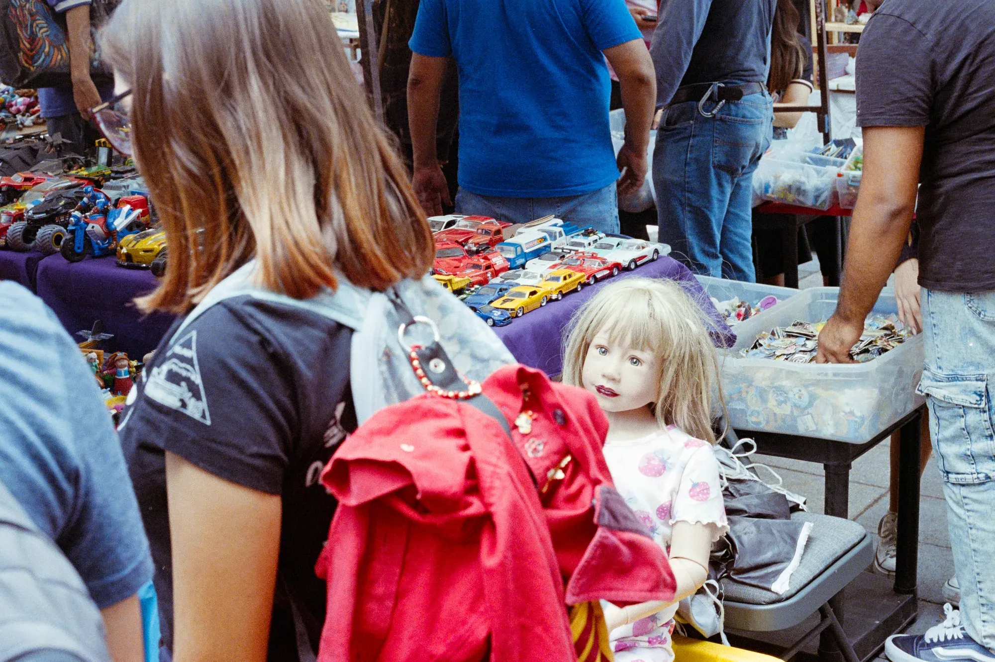 En un tianguis, una muñeca rubia observa desde un puesto mientras una niña pasa de espaldas