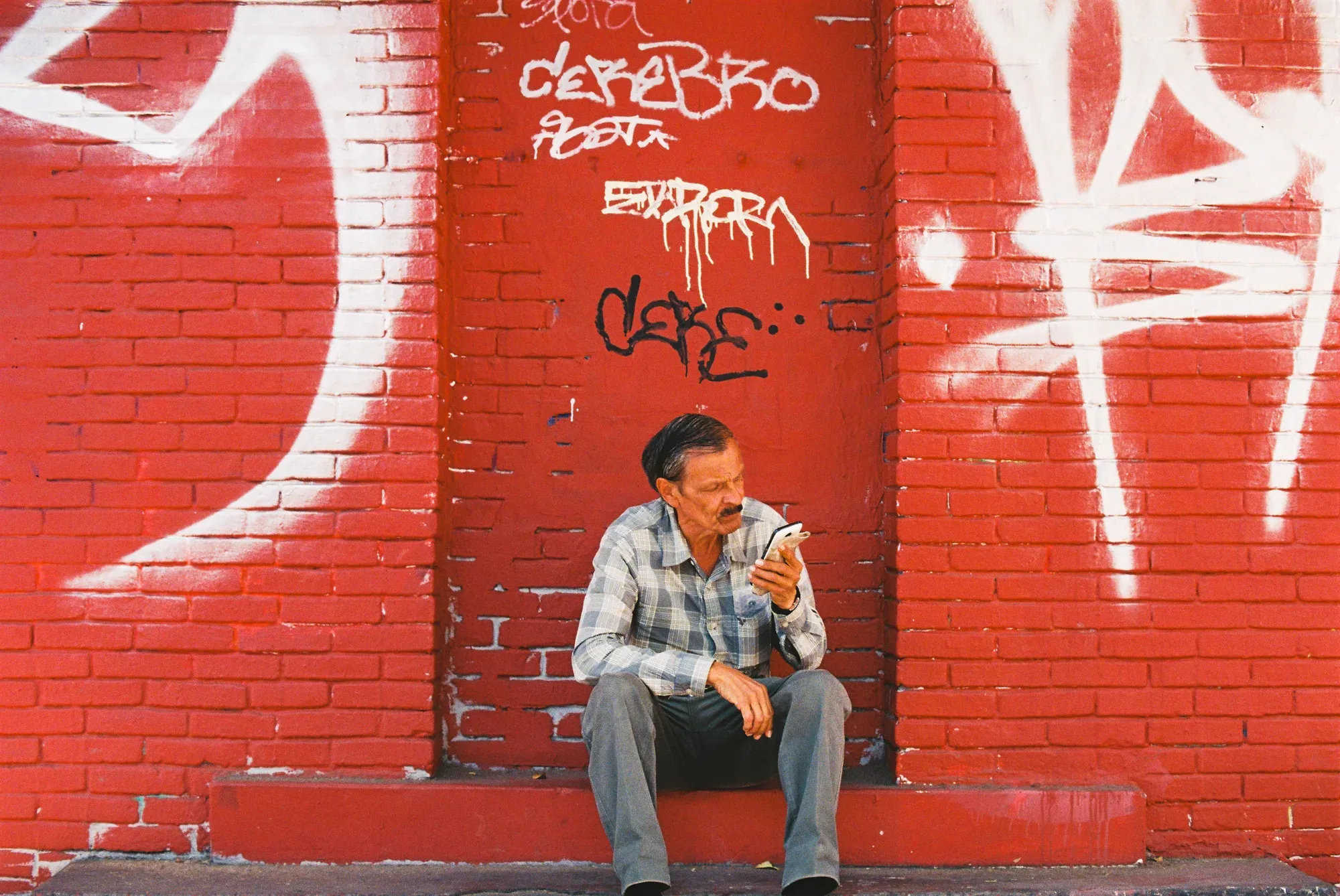 Hombre sentado frente a un muro rojo con grafiti mirando su celular